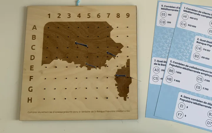 Banque Populaire Méditerranée : plateau en bois gravé à la carte du territoire, percé d’une grille de A à H et de 1 à 9 où sont plantés des drapeaux, à côté des fiches de quiz