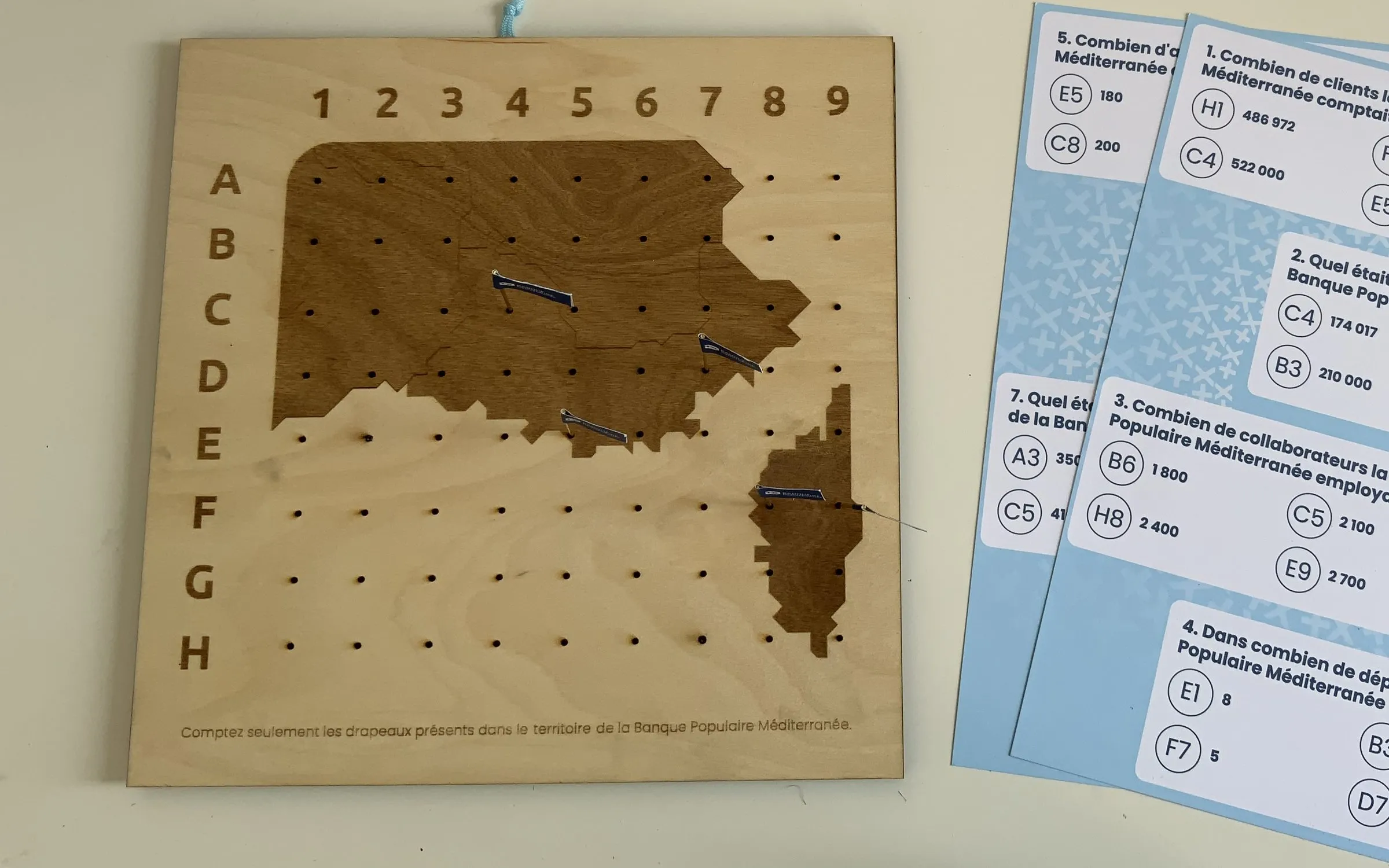 Banque Populaire Méditerranée : plateau en bois gravé à la carte du territoire, percé d’une grille de A à H et de 1 à 9 où sont plantés des drapeaux, à côté des fiches de quiz