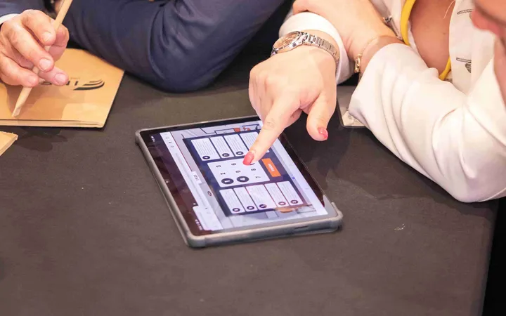 B.Talks 2024 : une participante tape un code sur le digicode affiché par la tablette, sur fond de bureaux virtuels, à côté d’une enveloppe kraft et d’un crayon