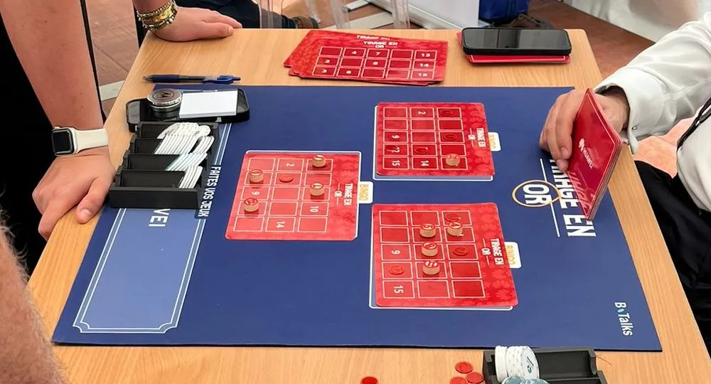 B.Talks 2025 : table Huawei du Grand Casino, grilles rouges « Tirage en or » garnies de pions sur le tapis bleu, porte-jetons et carte en main d’un participant