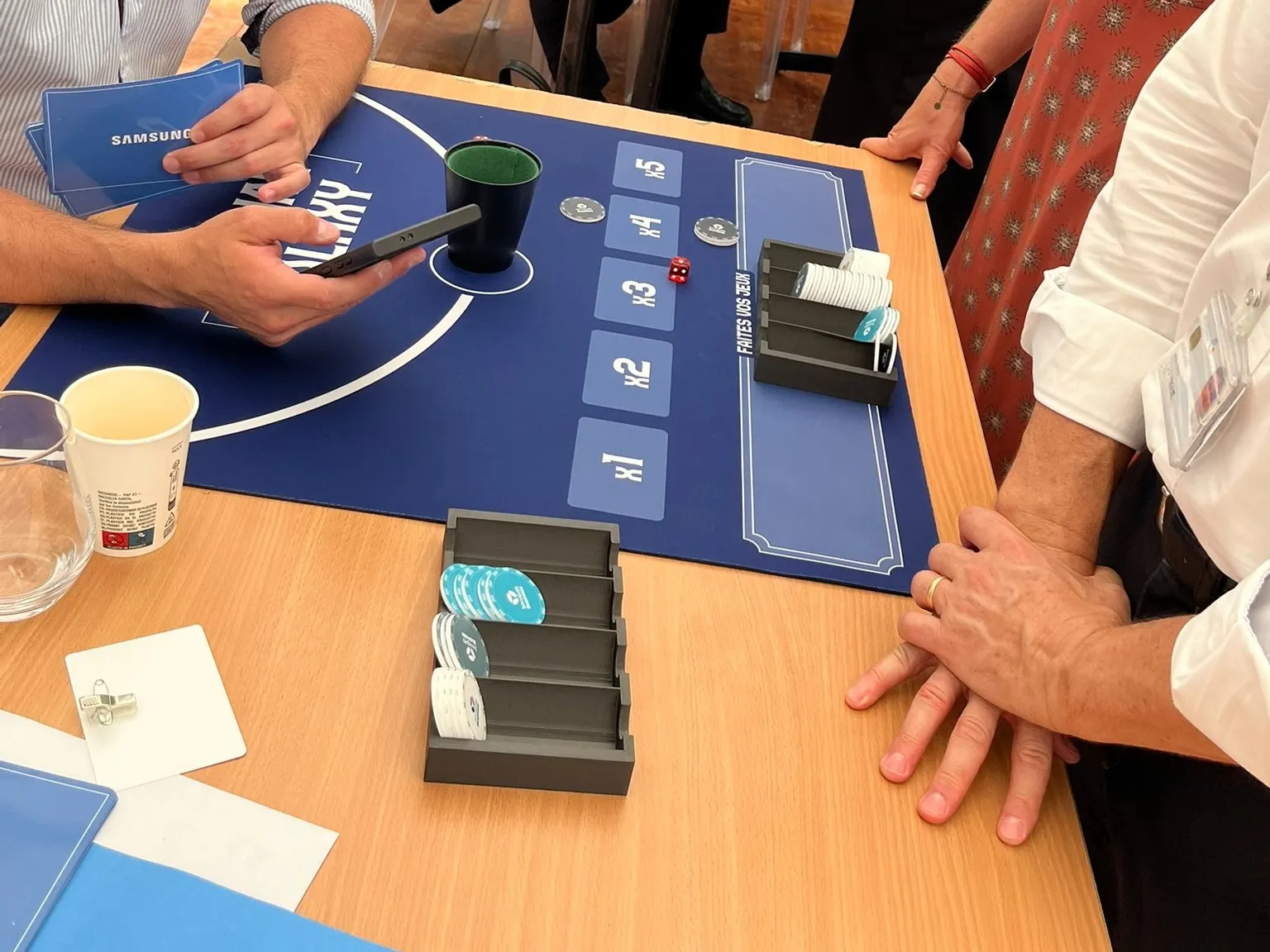 B.Talks 2025 : table Samsung du Grand Casino, tapis bleu « Cho-Han Galaxy » avec cases de mise x1 à x5, gobelet à dés, porte-jetons et cartes Samsung en main d’un participant