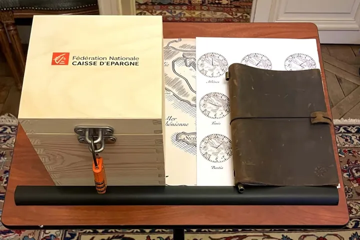 Caisse d’Épargne : coffret en bois « Fédération Nationale Caisse d’Epargne » fermé par un cadenas, carte maritime, planche d’horloges (Athènes, Tunis, Bastia) et carnet en cuir posés sur une table