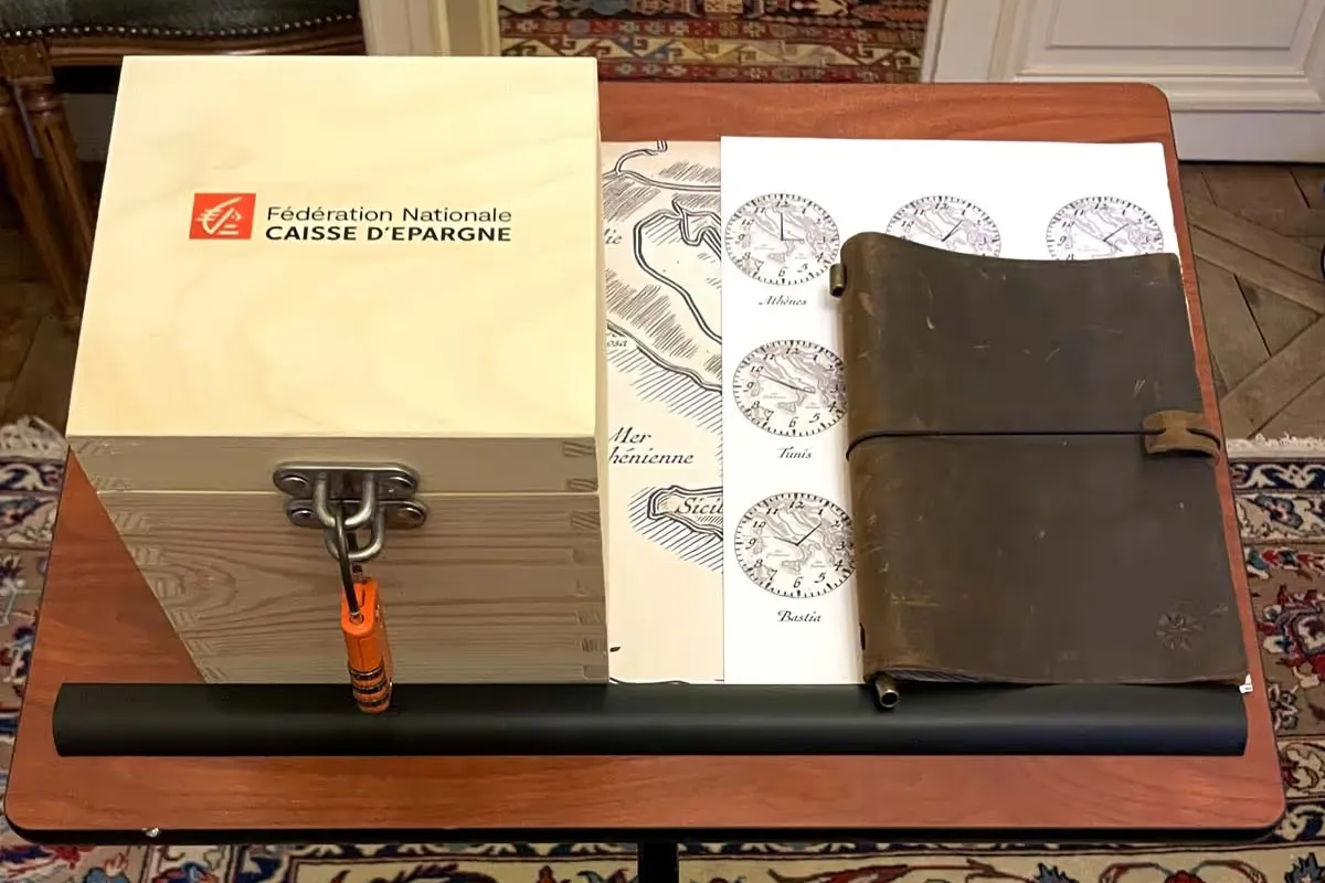 Caisse d’Épargne : coffret en bois « Fédération Nationale Caisse d’Epargne » fermé par un cadenas, carte maritime, planche d’horloges (Athènes, Tunis, Bastia) et carnet en cuir posés sur une table