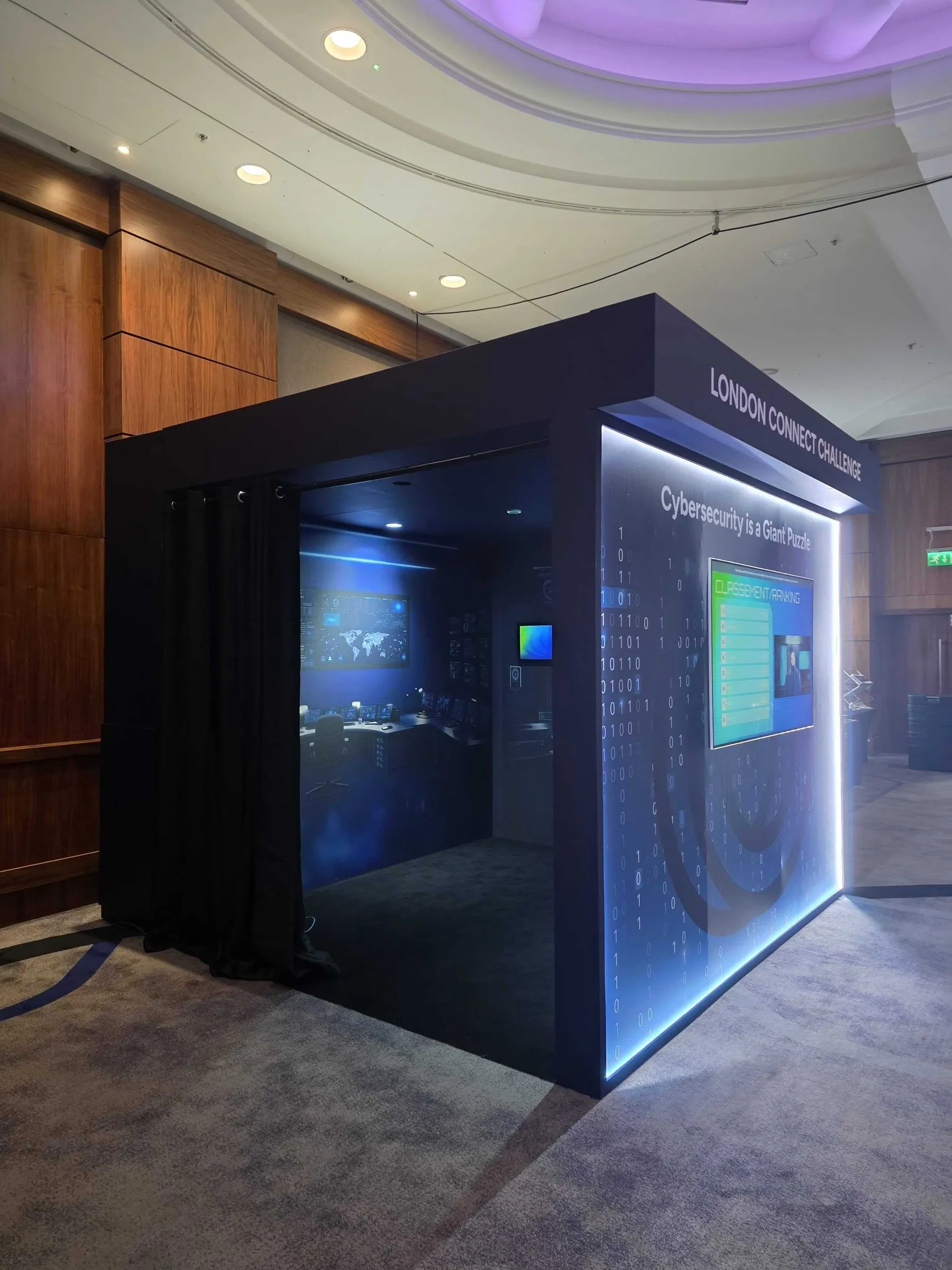 Distech Controls : la cabine « Cybersecurity is a Giant Puzzle » du London Connect Challenge, éclairée en bleu, avec un poste de contrôle à l’intérieur et le classement des équipes en façade