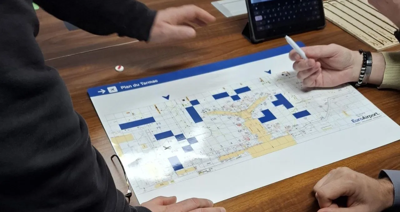 Serious game de gestion de crise EuroAirport : mains de l’équipe autour du plateau « Plan du Tarmac » couvert de pièces bleues, avec une tablette
