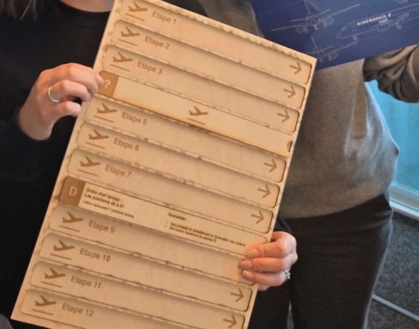 EuroAirport : planche en bois gravée des étapes 1 à 12 et plaque bleue « Air France E90 » à plans d'avion, deux éléments du matériel du serious game