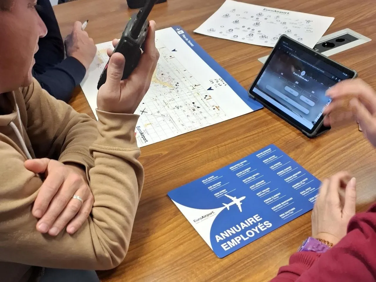 EuroAirport : un participant parle dans un talkie-walkie à côté du plan du tarmac, d’une tablette et de l’annuaire fictif des employés