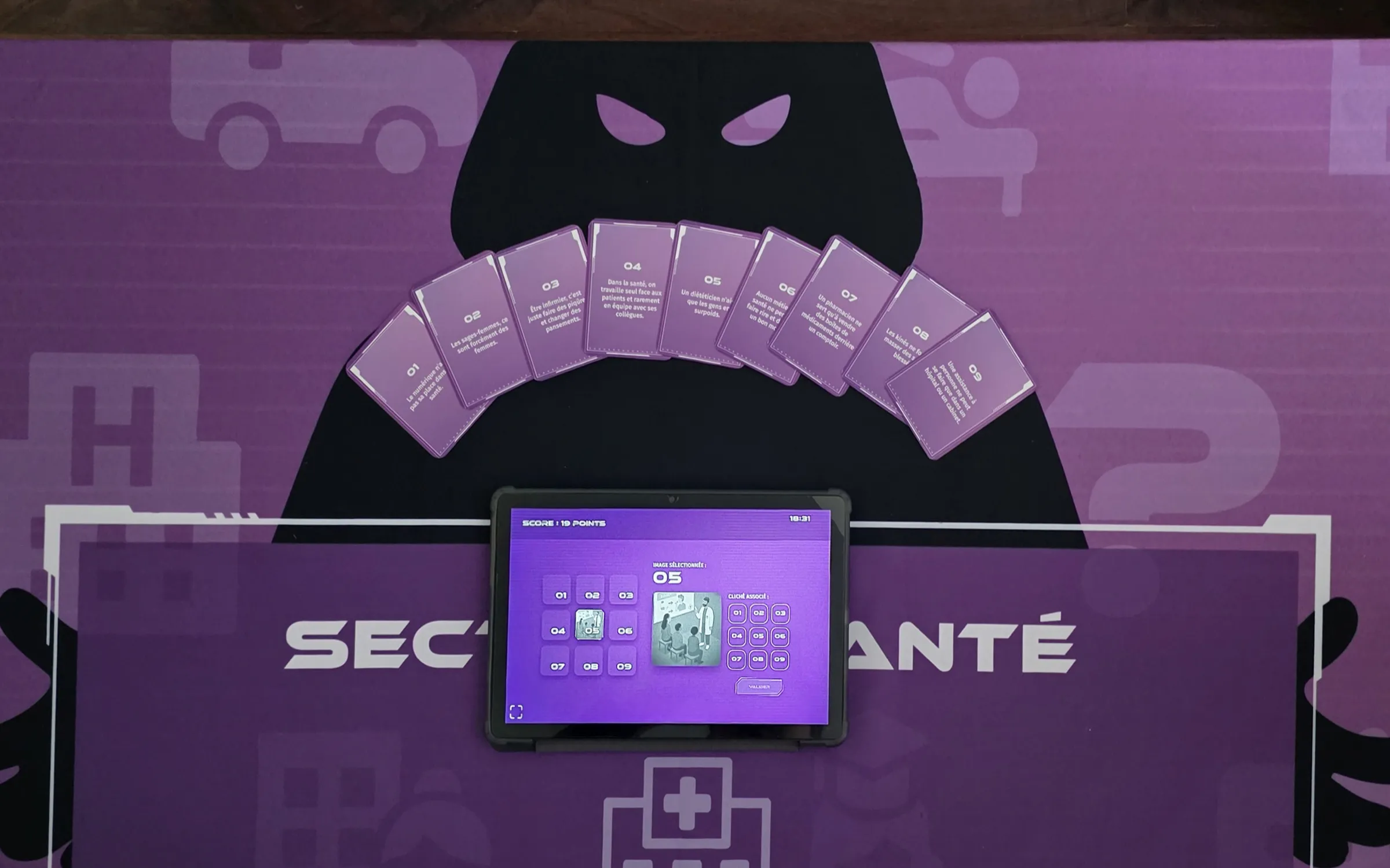 Béthune-Bruay : tapis de jeu « Secteur santé » vu du dessus, avec la silhouette encapuchonnée, l’éventail de cartes numérotées et la tablette du jeu